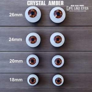 May include: Four pairs of round, white-based prosthetic eyes with amber-coloured irises and black pupils, labelled Crystal Amber. Sizes include 26mm, 24mm, 20mm, and 18mm. The text Life Like Eyes Dead Head Props is visible at the top right corner.