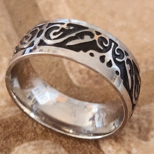 Può includere: Un anello d'argento con un designo nero intricato. L'anello è largo e ha una finitura liscia e lucida.