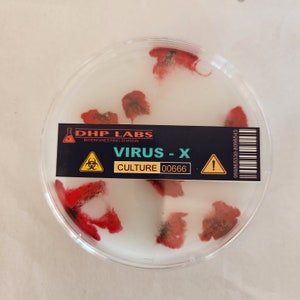 Puede incluir: Una placa de Petri con manchas rojas y negras sobre un fondo blanco. Una etiqueta dice "DHP LABS BIOENGINEERING DIVISION VIRUS - X CULTURE 00666" con un símbolo de riesgo biológico y una señal de advertencia.