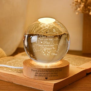 May include: A clear glass sphere lamp resting on a circular wooden base. The sphere is engraved with the text "Never forget you are the piece that made the difference" and "Olivia". The wooden base features the engraved text "Thank you for the work you did the person you are and the difference you made".