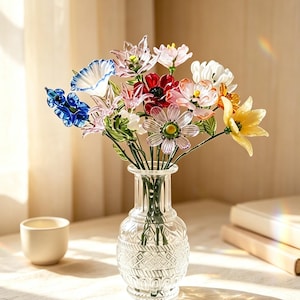 May include: A clear glass vase with a textured, diamond-patterned base holds a bouquet of multi-coloured glass flowers. The arrangement features various blooms in shades of blue, red, pink, white, and yellow, each with green glass stems and leaves.