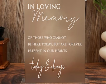 Op de afbeelding: Een helder acrylbord met de woorden "IN LOVING Memory" in witte letters. Het bord bevat ook de tekst "OF THOSE WHO CANNOT BE HERE TODAY, BUT ARE FOREVER PRESENT IN OUR HEARTS today & always."