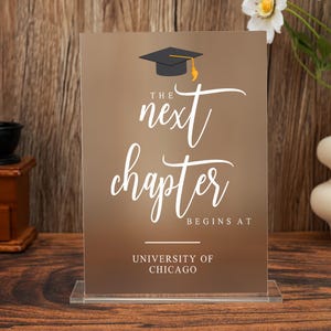 Könnte beinhalten: Ein Abschluss-Themenschild mit dem Text "The Next Chapter Begins At University of Chicago". Das Schild zeigt eine Grafik einer Abschlusskappe und wird in einem transparenten Acrylhalter präsentiert. Der Hintergrund ist ein brauner Farbverlauf.