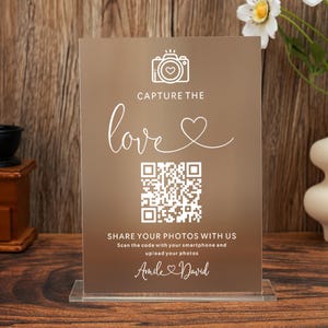 Puede incluir: Un letrero de boda de acrílico transparente con un fondo marrón. El letrero tiene texto blanco que dice "CAPTURE THE love" y "SHARE YOUR PHOTOS WITH US". También se incluye un código QR, junto con los nombres "Amile & David".