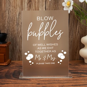 May include: A clear acrylic sign with the words "BLOW bubbles" in a cursive font. The sign also reads "OF WELL WISHES AS WE EXIT TOGETHER AS Mr. & Mrs. PLEASE TAKE ONE". The sign is on a clear stand.
