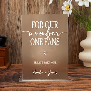 May include: A clear acrylic sign with the text "FOR OUR number ONE FANS" in white script. Below the text is a heart symbol, followed by "PLEASE TAKE ONE" and the names "Amelia + James". The sign is on a wooden surface.