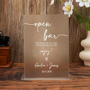 May include: A rectangular bronze-colored acrylic sign with a clear base. The sign features white text that reads: open bar, THE DRINKS ARE ON US BUT THE HANGOVER IS ON YOU, enjoy!, a heart symbol, Amelia + James, and the date 20.01.2026.