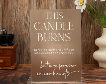 May include: A rectangular, bronze-colored sign with the text "THIS CANDLE BURNS" in white. Below, it reads "in loving memory of those who cannot be here today but are forever in our hearts." The sign is displayed on a clear acrylic base.