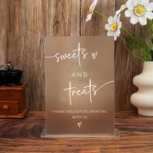 May include: A clear acrylic sign with the words "sweets and treats" in white script, along with "Thank you for celebrating with us." The sign is displayed on a wooden surface, with a coffee grinder and flowers in the background.