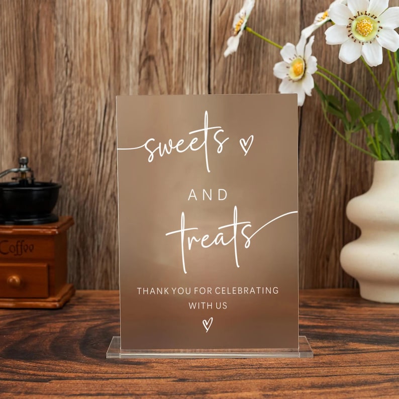 Op de afbeelding: Een helder acrylbord met de woorden "sweets and treats" in witte letters, samen met "Thank you for celebrating with us". Het bord staat op een houten oppervlak, met een koffiemolen en bloemen op de achtergrond.