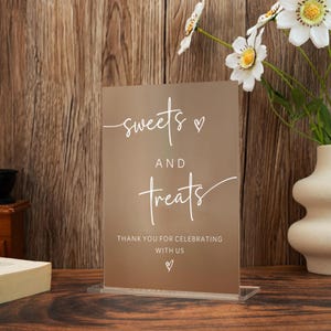 Op de afbeelding: Een helder acrylbord met de woorden "sweets and treats" in wit script. Het bord bevat ook de tekst "Thank you for celebrating with us" en een hartsymbool. Het bord wordt weergegeven op een houten oppervlak.