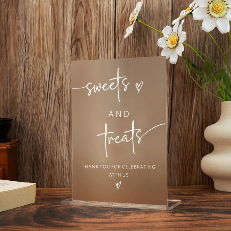 Op de afbeelding: Een helder acrylbord met de woorden "sweets and treats" in wit script. Het bord bevat ook de tekst "Thank you for celebrating with us" en een hartsymbool. Het bord wordt weergegeven op een houten oppervlak.