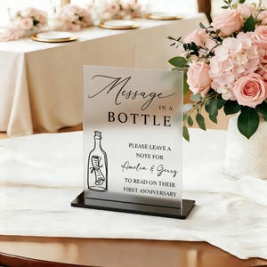 以下が含まれることがあります： 「Message in a Bottle」の文字とボトルのイラストが描かれた透明なアクリルサイン。サインには「Amelia & Gerryへ、結婚一周年のお祝いメッセージを書いてください」というテキストが含まれています。サインは黒いベースの上にあります。