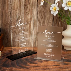 May include: Two bar menu signs. One is clear acrylic with a black base, the other is brown with a clear base. Both signs have the word "bar" in cursive, followed by a menu of drinks, names, and a date.