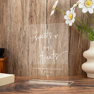 Op de afbeelding: Transparant acrylbord met de woorden "sweets and treats" in elegant wit schrift. Het bord bevat de zin "Thank you for celebrating with us" en een hartsymbool. Het bord staat op een houten oppervlak.