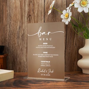 May include: A clear acrylic bar menu with a brown background. The menu lists beer, wine, and cocktails, with specific drink names. The names "Rachel & Jack" and the date "07.18.2025" are also displayed in white script.