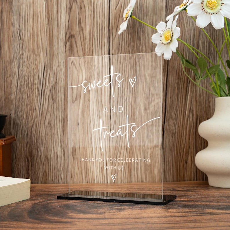 Op de afbeelding: Transparant acrylbord met de woorden "sweets and treats" in elegant wit schrift, perfect voor een desserttafel. Het bord bevat de zin "Thank you for celebrating with us" en een hartsymbool. Het staat op een zwarte basis.