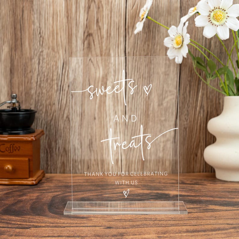 Op de afbeelding: Transparant acryl bord met de woorden "sweets and treats" in elegant wit schrift. Het bord bevat ook de tekst "Thank you for celebrating with us" en een hartsymbool. Perfect voor een bruiloft of feest.