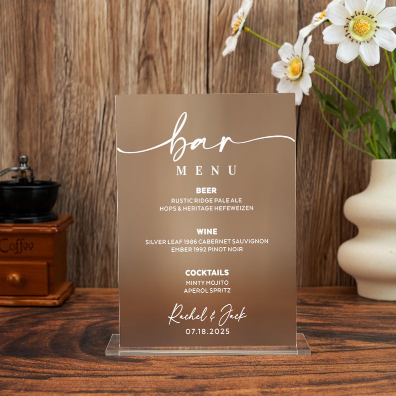 May include: A clear acrylic bar menu with a brown background. The menu lists beer, wine, and cocktails, with the names of the drinks. The top of the menu says "bar" in cursive. The bottom of the menu says "Rachel & Jack" and the date "07.18.2025".
