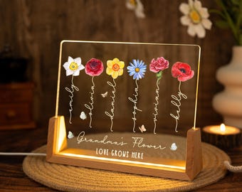 May include: A personalized LED night light with a wooden base. The clear acrylic panel features colorful flower illustrations and names: Emily, Olivia, Daniel, Sophia, Vivian, and Kelly. The text "Grandma's Flower" and "LOVE GROWS HERE" is also displayed.
