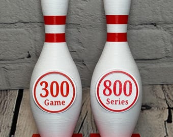 May include: Two white bowling pin trophies with red accents. One pin reads "300 Game" and the other "800 Series." Each sits on a red base with the text "Your Name Here" and the date 12-12-26.