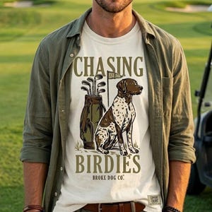 May include: An off-white t-shirt featuring a graphic of a brown and white spotted dog sitting next to a golf bag with clubs and a flag. The text reads CHASING BIRDIES and BROKE DOG CO. The shirt is worn under an olive green button-down shirt.