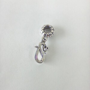 Könnte beinhalten: Ein silberfarbener Metall-Charm-Anhänger mit einer hängenden Schlangenfigur. Die Perle verfügt über eine kreisförmige, texturierte Öse zur Befestigung, während die Schlange in einer gewundenen Position mit glatter, polierter Oberfläche dargestellt ist.