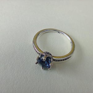 May include: A silver ring with a large, dark blue gemstone at the center. The band is detailed with small, dark stones. The ring is set against a light background, highlighting its design and color.