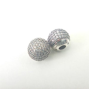 May include: Two spherical silver charms covered in small, clear pavé-set stones. One charm shows the word PANDORA engraved on its smooth, polished silver side. The charms have a metallic finish and a textured, sparkling surface from the stones.