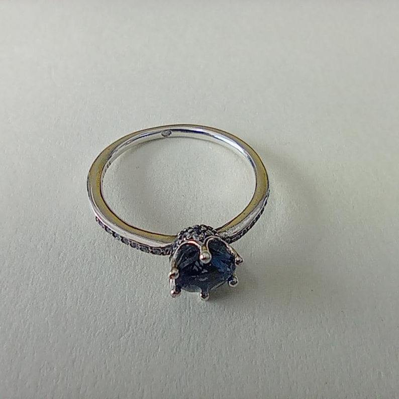 May include: A silver ring with a large, dark blue gemstone at the center. The band is thin and has small, dark accents. The gemstone is surrounded by a crown-like setting. A piece of jewelry.