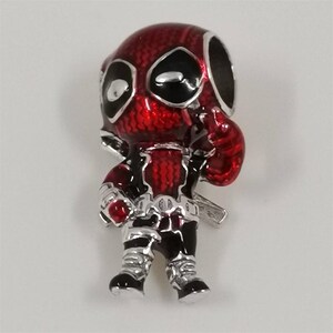 Op de afbeelding: Een rode en zwarte Deadpool-bedel met zilveren accenten. De bedel toont het personage in een staande pose, met een hand omhoog. De bedel is waarschijnlijk gemaakt van metaal en emaille.