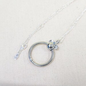 Op de afbeelding: Een zilveren ketting met een cirkelvormige, getextureerde hanger en een bloemvormige sluiting. De hanger is bevestigd aan een fijne ketting. De ketting wordt gepresenteerd op een lichte achtergrond.