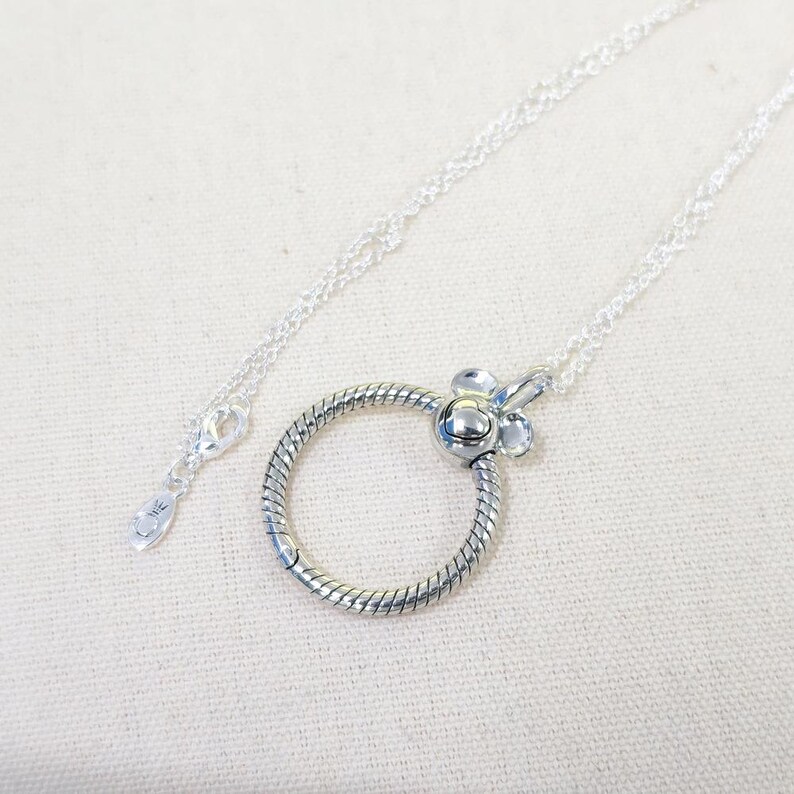 Op de afbeelding: Een zilveren ketting met een cirkelvormige, getextureerde hanger en een bloemvormige sluiting. De hanger is bevestigd aan een fijne ketting. De ketting wordt gepresenteerd op een lichte achtergrond.