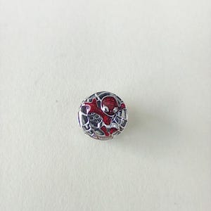May include: A circular silver charm with a red and black Spider-Man design. The charm has a web pattern around the edges and a red Spider-Man figure in the centre. The charm is likely made of metal.