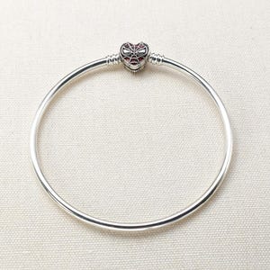Könnte beinhalten: Ein silberfarbenes Armreif-Armband mit einem herzförmigen Anhänger. Der Anhänger hat ein rotes und schwarzes Spinnennetz-Design. Das Armband ist kreisförmig und hat eine glatte, polierte Oberfläche. Der Anhänger ist mittig auf dem Armband.
