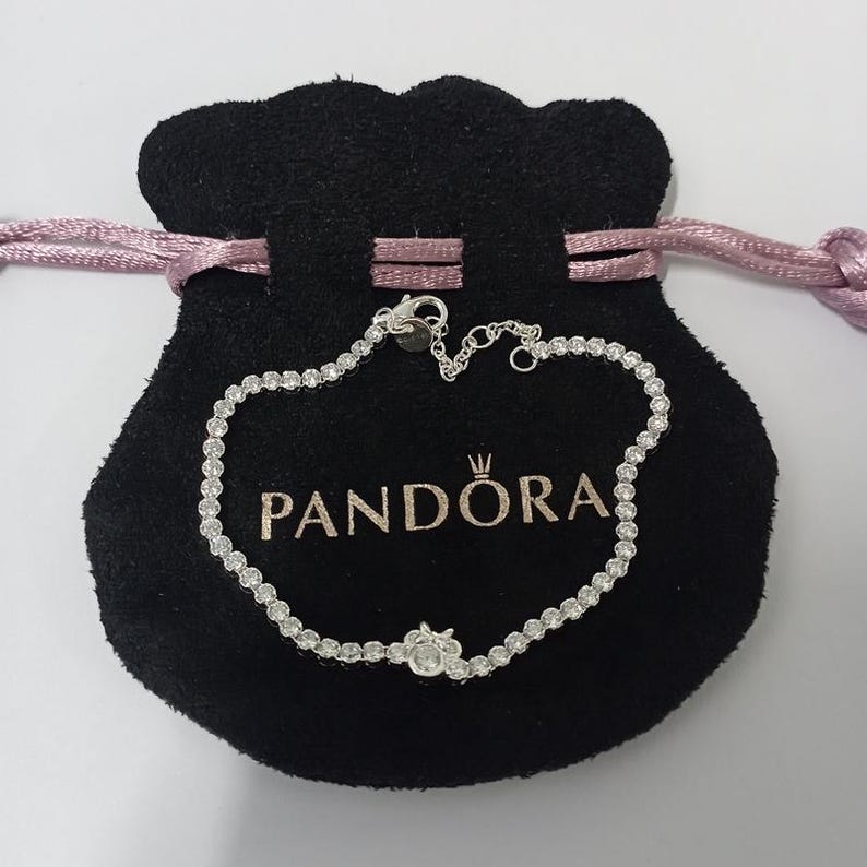 May include: A silver bracelet with numerous small, clear stones is displayed on a black velvet pouch. The pouch has the brand name "PANDORA" printed in white. The bracelet is delicate and features a floral design.