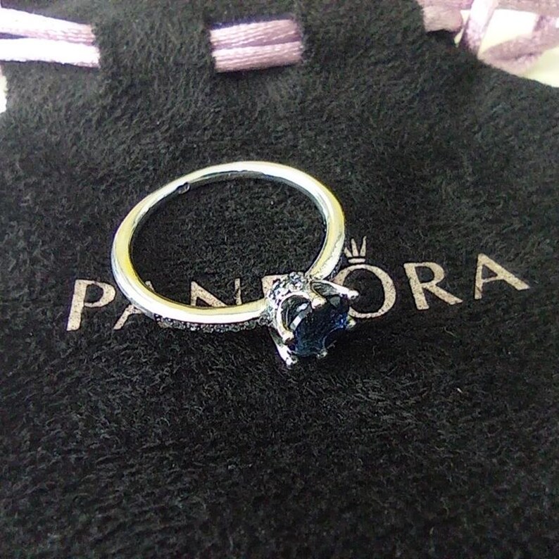 May include: A silver ring with a large, dark blue gemstone. The ring is set on a black velvet pouch with the word "PANDORA" printed on it. The ring has a delicate band and small accent stones.