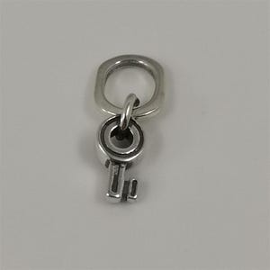 May include: A silver-colored key charm with a small key design. The charm is attached to a larger, oval-shaped ring. The key has a detailed, circular head and a classic key shape. The metal has a polished finish.