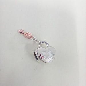 May include: A silver heart-shaped charm with a key pendant. The charm features a design of two figures kissing, with red accents. The key is rose-gold colored. The charm is likely for a bracelet or necklace.