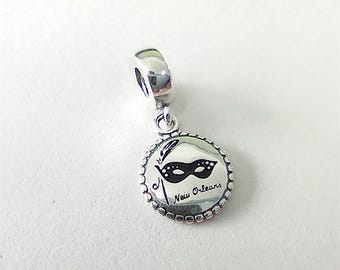 Peut inclure: Un charm pendant en argent composé d'un disque circulaire à bord perlé. Le centre du disque présente un motif de masque de carnaval noir avec une plume et l'inscription New Orleans écrite en écriture cursive sous le masque.