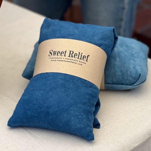 May include: Two blue fabric pillows are wrapped with a brown paper band that reads "Sweet Relief" with the website address. The pillows are on a white surface. The pillows are a deep blue color.