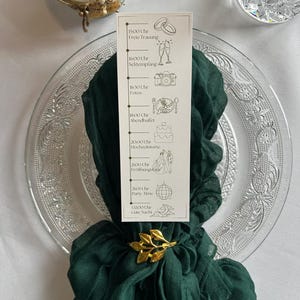Könnte beinhalten: Eine Hochzeitstafel mit einem dunkelgrünen Schal, einem goldenen Blätter-Serviettenring und einer Karte mit einem deutschen Hochzeitsablauf. Eine Kerze und ein Kristallglas sind ebenfalls zu sehen.