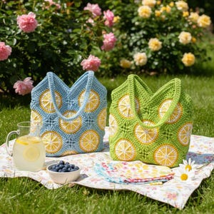 May include: Two crochet tote bags, one light blue and one green, each adorned with yellow and white lemon slice designs. The bags sit on a floral picnic blanket with a pitcher of lemonade and a bowl of blueberries.