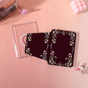 May include: A clear phone case with a white ring grip and two burgundy cardholders. The cardholders feature a floral and crescent moon design in gold and white. A small pink pig figurine and a white cat figurine are also present.