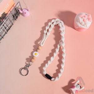 May include: A cream-colored macrame lanyard with a daisy charm and wooden beads. The lanyard has a silver-colored clasp and a black accent piece. The background is a light pink surface with decorative items.