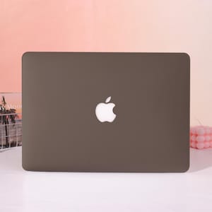 May include: A dark gray laptop with a white Apple logo centered on the lid. The laptop is closed and appears to be on a white surface. The background is a gradient of pink and peach.