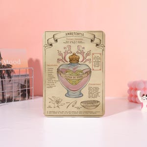 May include: A decorative card featuring a potion bottle design. The bottle is heart-shaped, filled with pink liquid, and labeled "FILTRO DE AMOR." The card has a vintage aesthetic with text in Spanish and illustrations of ingredients.