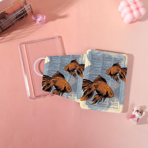 May include: Clear phone case and two phone inserts with a goldfish design. The inserts feature a blue background with dictionary text and orange goldfish illustrations. The case is transparent, and the inserts are rectangular.