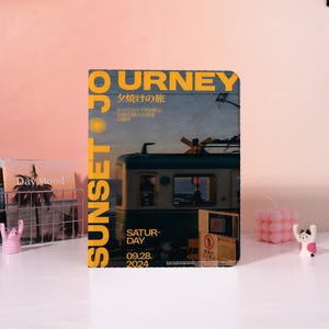 Könnte beinhalten: Ein Notizbuch mit dem Wort "JOURNEY" in Gelb und dem Wort "SUNSET" in Gelb vertikal. Das Notizbuch zeigt ein Bild eines Zuges und das Datum "09.28. 2024".