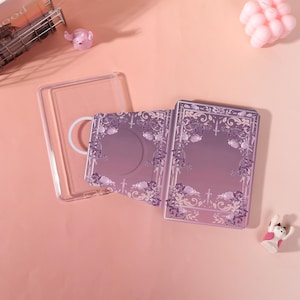May include: A clear phone case and two purple and white tarot card decks with floral and sword designs are displayed on a pink surface. The cards have a gradient background and decorative borders. A small pink pig figurine and a white cat figurine are also present.